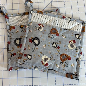 May include: Two quilted pot holders with a light blue background. The fabric is patterned with cartoon chickens in various colors and small white flowers. The top edge has a striped pattern and a loop for hanging.