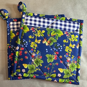 May include: Two blue quilted pot holders with a vibrant berry and butterfly pattern. The design includes red raspberries, blueberries, and green leaves on a dark blue background. Each pot holder has a white and blue checkered trim and a loop for hanging.
