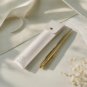 May include: A gold pen and a white leather pen case. The pen case has a snap closure and the brand name "mumiikart" is embossed on the front. The pen is next to the case on a white surface. The pen is a classic design.
