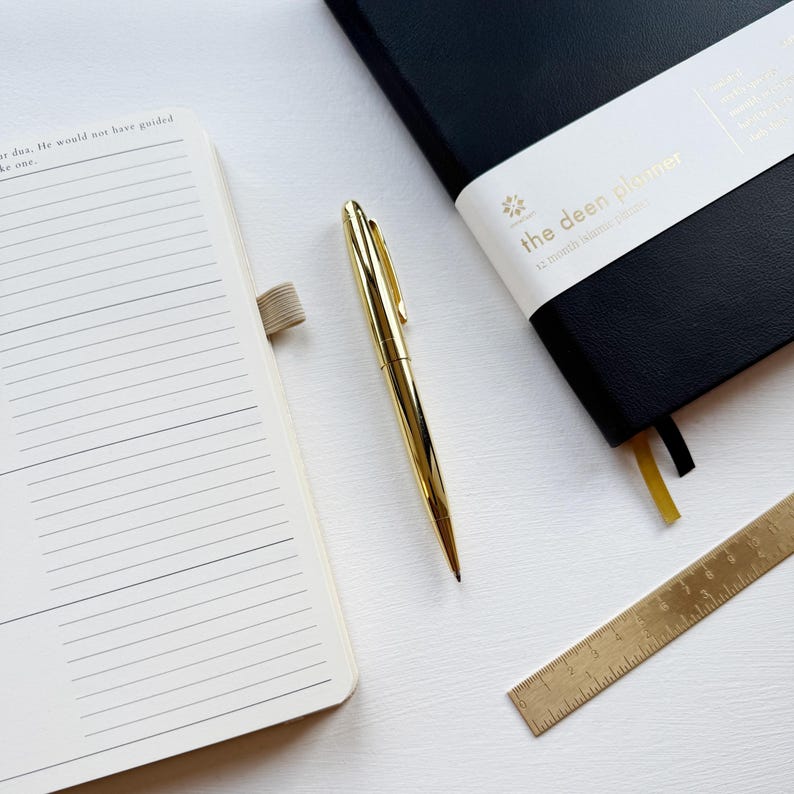 May include: A gold pen, a black planner with the text "the deen planner", a notebook, and a brass ruler are arranged on a white surface. The planner has a white band with gold text and a yellow ribbon bookmark.
