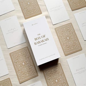 Könnte beinhalten: Eine weiße Box mit dem Text "THE BOX OF BARAKAH" und 52 islamischen Erinnerungen. Die Box ist umgeben von Karten mit beigefarbenen floralen Mustern und weißen Karten mit arabischem Text.