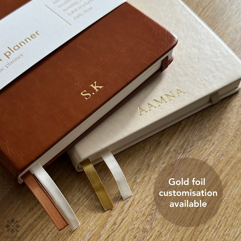 May include: Two leather-bound journals with gold foil lettering. The top journal is brown and says "S.K." The bottom journal is cream and says "AAMNA". Both journals have white ribbon bookmarks.