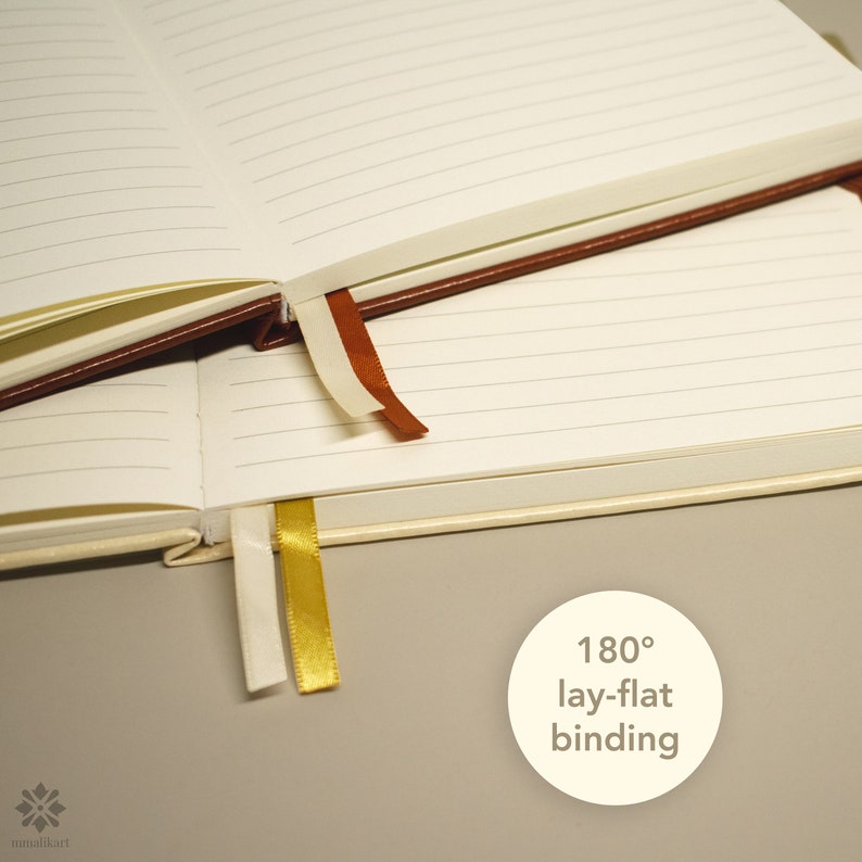 May include: A white notebook with brown and gold ribbon bookmarks. The notebook is open to show the lined pages. The text "180&deg; lay-flat binding" is printed on a white circle.