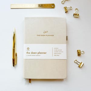 May include: A beige planner with gold text reading "THE DEEN PLANNER" and "the deen planner". A gold pen, ruler, and binder clips are arranged around the planner on a white surface. The planner includes undated weekly spreads and habit trackers.