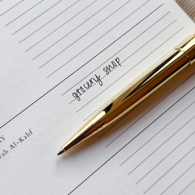 May include: A gold pen rests on lined paper, with the words "grocery shop" handwritten in black ink. The pen's surface is highly polished and reflective. The paper has a pale, neutral colour.