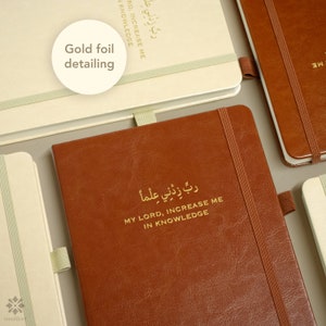 May include: Two leather-bound journals with gold foil detailing. The top journal is cream colored with the text "رب زدني على" in gold lettering. The bottom journal is brown with the text "ربِّ زِدْنِي عِلْماً" in gold lettering. The text translates to "My Lord, increase me in knowledge".