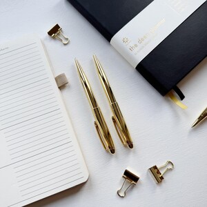 May include: Overhead view of stationery items: two gold pens, gold binder clips, a black journal with "the deen planner" text, and an open notebook with lined pages. The items are arranged on a white surface.