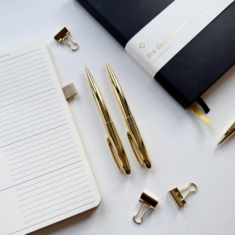 May include: Overhead view of stationery items: two gold pens, gold binder clips, a black journal with "the deen planner" text, and an open notebook with lined pages. The items are arranged on a white surface.