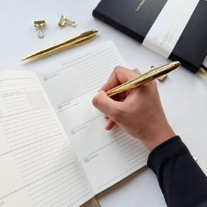 May include: A gold pen writing in an open notebook with a black cover. The notebook is open to a weekly planner page. Two gold binder clips and another gold pen are also visible.