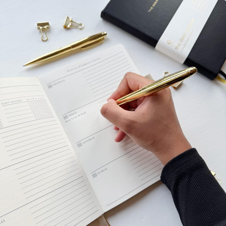 May include: A gold pen writing in an open notebook with a black cover. The notebook is open to a weekly planner page. Two gold binder clips and another gold pen are also visible.
