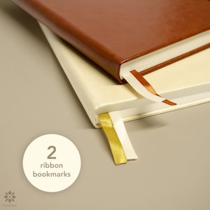 May include: Two notebooks with leather covers, one brown and one cream, stacked on top of each other. Both notebooks have ribbon bookmarks, one gold and one orange.