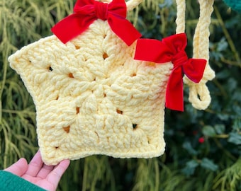 May include: A handmade, star-shaped crochet bag in a soft yellow hue, adorned with two vibrant red velvet bows. The bag features a textured stitch pattern and a matching yellow handle. Text on the image reads "Star Bag Pattern".