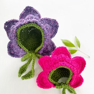 May include: Two handmade crochet flower hats. One is purple with green accents, and the other is hot pink with green accents. The hats are shaped like flowers with petals and green leaves and stems.