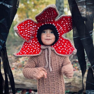 May include: A child in a handmade Demogorgon costume. The costume includes a large red and white flower-like headpiece, a black hood, and a brown crocheted jumpsuit. The costume is designed to resemble the creature from the popular science fiction series.