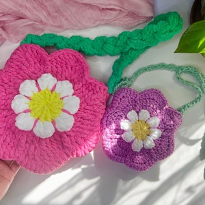 Può includere: Due borse a forma di fiore all'uncinetto, una rosa e una viola, con dettagli di fiori bianchi. La borsa rosa è più grande e ha una tracolla in crochet verde.
