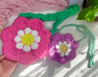 May include: Two crocheted flower-shaped purses, one pink and one purple, with white flower accents. The pink purse is larger and has a green crocheted strap.