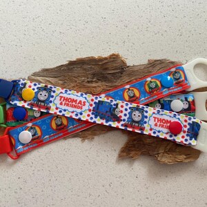 May include: Three Thomas & Friends themed dummy clips. Each clip features a colourful design with images of Thomas the Tank Engine and friends. The clips have different coloured clasps and buttons, and white loops for attaching dummies.