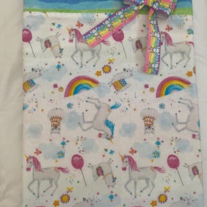 May include: A white blanket with a pattern of unicorns, rainbows, balloons, and clouds. A rainbow ribbon bow is attached to the blanket. The top of the blanket has a gradient of pink, blue, and purple.