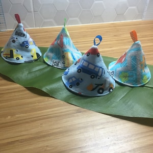 May include: Four light blue fabric bowl covers with a transport theme, featuring cars, trucks, and buses. Each cover has a loop at the top in red, green, blue, and orange. The covers are displayed on a green leaf.