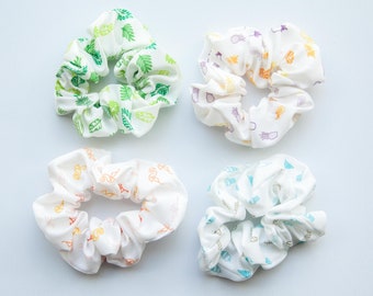 May include: Four white scrunchies with different colorful patterns. The patterns include green leaves, pineapples, and a rainbow design.