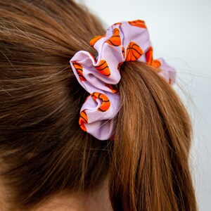 May include: A light purple scrunchie with a repeating pattern of orange and black basketballs. The scrunchie is tied around a ponytail of brown hair, creating a sporty and stylish hair accessory.