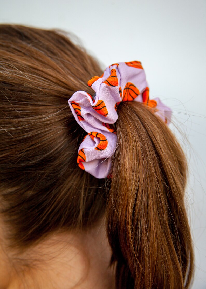 May include: A light purple scrunchie with a repeating pattern of orange and black basketballs. The scrunchie is tied around a ponytail of brown hair, creating a sporty and stylish hair accessory.