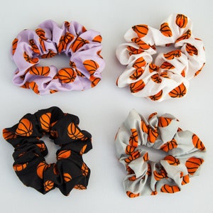 May include: Four scrunchies with basketball patterns. The scrunchies are in various colors: lavender, white, black, and gray. Each scrunchie features a repeating orange basketball design. These hair accessories are perfect for sports fans.