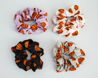 May include: Four scrunchies with basketball patterns. The scrunchies are in various colors: lavender, white, black, and gray. Each scrunchie features a repeating orange basketball design. These hair accessories are perfect for sports fans.