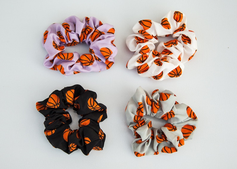 May include: Four scrunchies with basketball patterns. The scrunchies are in various colors: lavender, white, black, and gray. Each scrunchie features a repeating orange basketball design. These hair accessories are perfect for sports fans.
