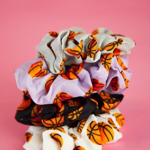 May include: A stack of four scrunchies with a basketball print. The scrunchies are in various colors, including white, black, lavender, and light gray. The basketballs are orange and black.