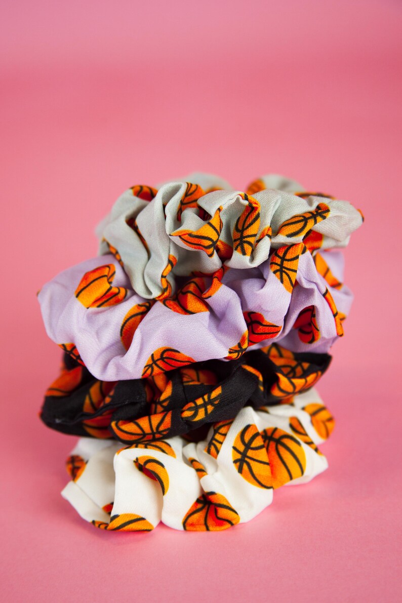 May include: A stack of four scrunchies with a basketball print. The scrunchies are in various colors, including white, black, lavender, and light gray. The basketballs are orange and black.