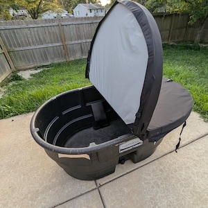 May include: A black Rubbermaid stock tank with a black and white cover. The tank is oval-shaped and sits on a concrete surface, with a grassy background and a wooden fence.