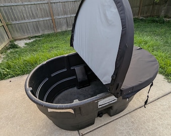 May include: A black Rubbermaid stock tank with a black and white cover. The tank is oval-shaped and sits on a concrete surface, with a grassy background and a wooden fence.