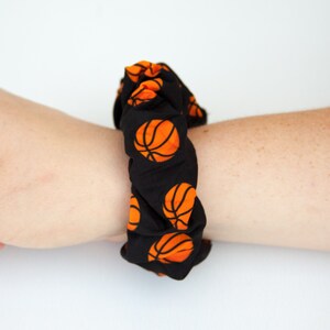 May include: Black scrunchie with a repeating pattern of orange basketballs. The scrunchie is wrapped around a wrist, showcasing the fabric's design and texture. This accessory is suitable for sports enthusiasts.