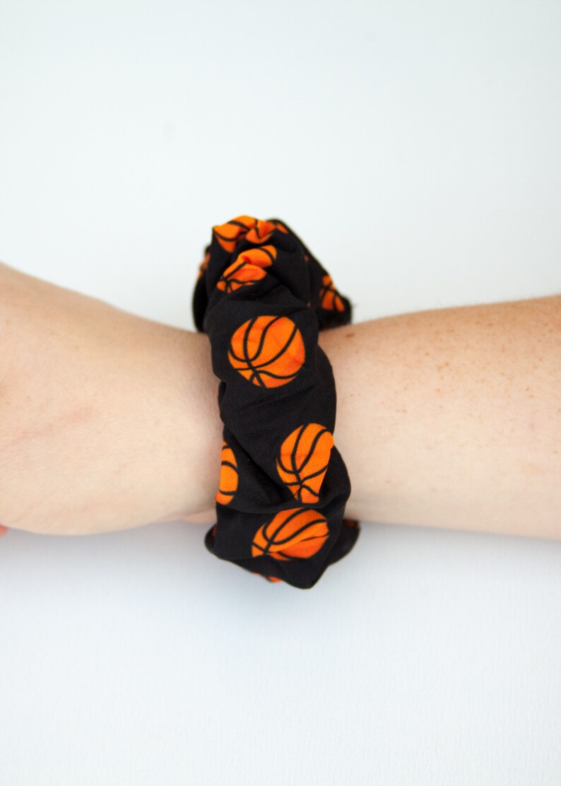 May include: Black scrunchie with a repeating pattern of orange basketballs. The scrunchie is wrapped around a wrist, showcasing the fabric's design and texture. This accessory is suitable for sports enthusiasts.