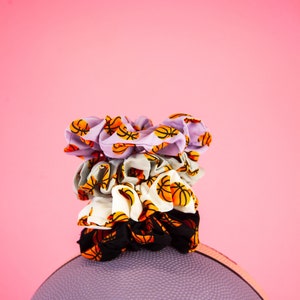 May include: A stack of four scrunchies with basketball patterns in various colors, including lavender, gray, white, and black, sits atop a purple basketball. The scrunchies feature orange and black basketball graphics. The background is a solid pink.