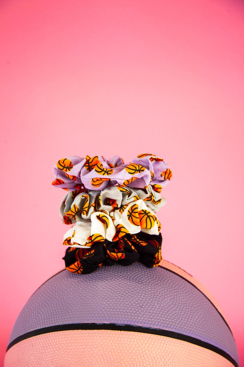 May include: A stack of four scrunchies with basketball patterns in various colors, including lavender, gray, white, and black, sits atop a purple basketball. The scrunchies feature orange and black basketball graphics. The background is a solid pink.