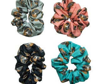 May include: Four scrunchies in various colors: light blue, coral, black, and teal. Each scrunchie features a repeating pattern of a dog's face in brown, black, and white. These fabric hair accessories are perfect for adding a touch of fun to any hairstyle.