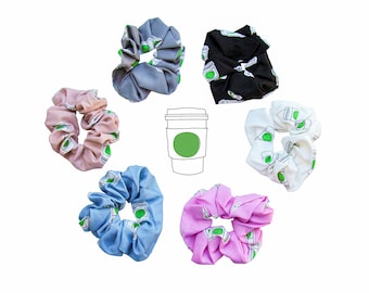 May include: Six colorful scrunchies with a coffee cup print. The scrunchies are in shades of pink, blue, gray, white, and black. The coffee cup is outlined in black and has a green lid and a green logo.