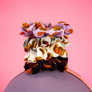 May include: A stack of scrunchies with a basketball pattern in various colors, including lavender, gray, white, and black, sits atop a purple basketball. The scrunchies feature an orange and black basketball design.