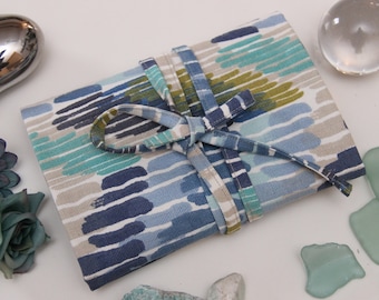 May include: A blue and white fabric pouch with a green and yellow pattern. The pouch is tied with a blue ribbon and is surrounded by sea glass and decorative stones.