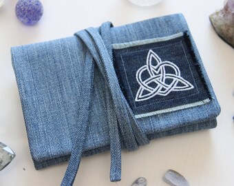 Puede incluir: Una bolsa de mezclilla azul con un diseño de nudo celta blanco bordado en un parche azul oscuro. La bolsa tiene un cierre de corbata y está rodeada de varios cristales y piedras.