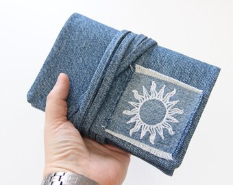 Puede incluir: Un cuaderno de mezclilla azul con un diseño de sol bordado en blanco. El cuaderno está envuelto con una correa de mezclilla azul.