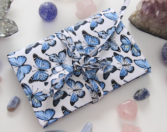 May include: A white fabric journal with a blue butterfly pattern and a white ribbon tied around it. The journal is surrounded by various crystals and stones.