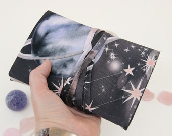 May include: A black fabric journal with a celestial design featuring a watercolor planet and stars. The journal is tied closed with a pink ribbon.