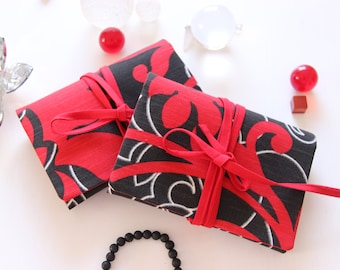May include: Two small red and black fabric pouches with a white floral design. The pouches are tied with red ribbon and are sitting on a white surface.