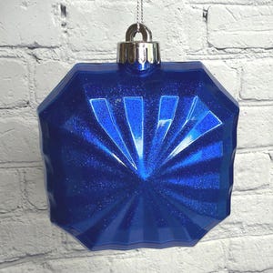 May include: A faceted, geometric blue Christmas ornament with a silver metal cap and hanging string. The ornament has a glittery finish and is suspended against a white brick wall.
