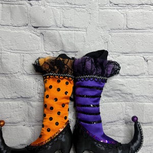 May include: A pair of decorative witch boots featuring one orange boot with black polka dots and one purple boot with black horizontal stripes. Both boots have black curled toes with glittery pom-poms, black heels, and black lace trim at the top.