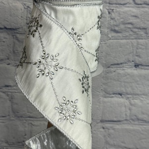 May include: A roll of white ribbon with silver snowflake embellishments. The ribbon has a satin finish and is trimmed with silver edging. The snowflakes are made of clear, sparkling rhinestones, adding a touch of elegance. Ideal for holiday decorations and crafts.