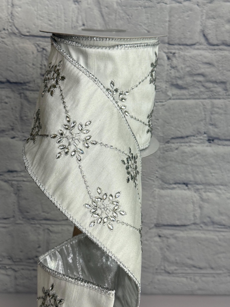 May include: A roll of white ribbon with silver snowflake embellishments. The ribbon has a satin finish and is trimmed with silver edging. The snowflakes are made of clear, sparkling rhinestones, adding a touch of elegance. Ideal for holiday decorations and crafts.